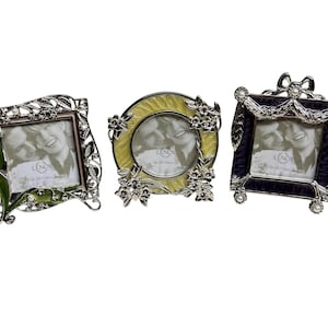 May include: Three decorative picture frames with metallic silver finishes. The left frame is square with green enamel leaves. The center frame is circular with yellow enamel and floral accents. The right frame is square with purple enamel and a bow.