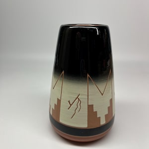 May include: A ceramic vase with a black top that fades to a cream color, featuring a geometric design in brown and cream. The vase has a reddish-brown base and a black band near the bottom. The design resembles a mountain range.