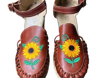 May include: A pair of brown leather platform sandals with a woven design and ankle straps. Each sandal features an embroidered sunflower with yellow petals and a green stem. The heels are approximately 3 inches high.
