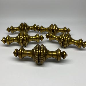 May include: Six antique brass door knobs with a detailed, ornate design. The knobs are a warm gold color and have a classic, vintage aesthetic. They are likely for a cabinet or drawer.
