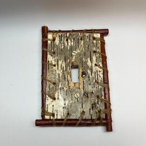 May include: A light switch cover made from birch bark with a wooden frame. The frame is made of thin branches that are woven together.