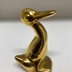 May include: A small, gold-coloured ceramic duck figurine. The duck is sitting with its head tilted to the side and its beak slightly open.