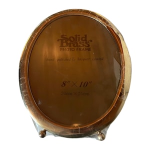 May include: Oval solid brass photo frame, hand polished and lacquer coated. The frame is 8" x 10" and features the text "Solid Brass PHOTO FRAME". The frame is packaged in clear plastic.