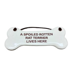 May include: A white, bone-shaped ceramic sign featuring dark brown text that reads A SPOILED ROTTEN RAT TERRIER LIVES HERE. The sign includes two small holes at the top with a thin, dark brown cord attached for hanging purposes.