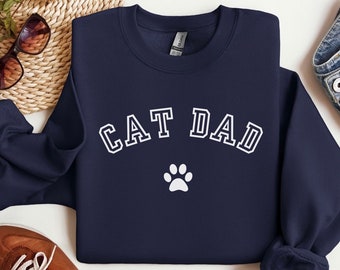 Może przedstawiać: Bordowy sweter z napisem "CAT DAD" w kolorze białym i białą odbitką łapy pod spodem.