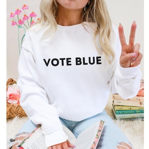 Puede incluir: Sudadera blanca con el texto "VOTE BLUE" impreso en negro.