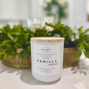 May include: A white candle with a wooden lid, labeled "Vanilla Patchouli" with "Wylie Candle Co." branding. The candle is 13 oz. and made with soy and coconut apricot creme wax. The background includes greenery and flowers.