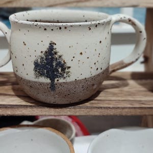 May include: Handmade ceramic mugs with a tree design. The mugs are white with brown speckles and a brown base. The tree is a dark blue colour. The mugs are on a wooden shelf with other ceramic bowls.