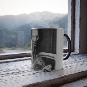 May include: A black and white heat-sensitive mug featuring a disturbing illustration of a pale, gaunt figure with a wide, toothy grin. The mug's handle is black, and the background is a window with a mountain view.