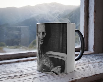 Può includere: Una tazza termosensibile in bianco e nero con una inquietante illustrazione di una figura pallida e smunta con un ampio sorriso dentuto. Il manico della tazza è nero e lo sfondo è una finestra con vista sulle montagne.