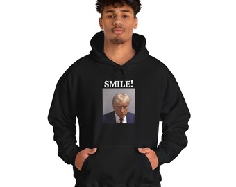 以下が含まれることがあります： 前面に男性の顔の白のグラフィックと「SMILE！」というテキストが印刷された黒いフード付きスウェットシャツです。