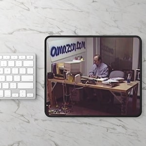 May include: A black and white photo of a man sitting at a desk in an office. The desk is cluttered with papers and computer equipment. The man is wearing a light blue shirt and is typing on a keyboard. The office has a sign on the wall that says "amazon.com".