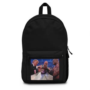 Könnte beinhalten: Schwarzer Rucksack mit einer Vordertasche und einem kleinen Bild eines Affen, der sich von Donald Trump und zwei anderen Männern die Haare schneiden lässt. Der Rucksack hat einen oberen Griff und einen Reißverschluss.