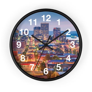 Puede incluir: Un reloj con marco negro con una imagen de ciudad. La esfera del reloj tiene números y manecillas blancas. La imagen de la ciudad muestra una ciudad de noche con edificios iluminados.