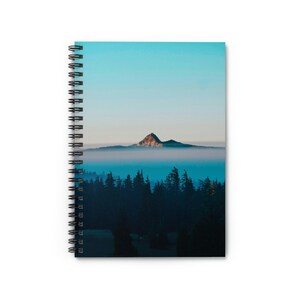 May include: A misty mountain peak rises above a forest of dark green trees against a blue sky. The fog creates a soft, ethereal effect.