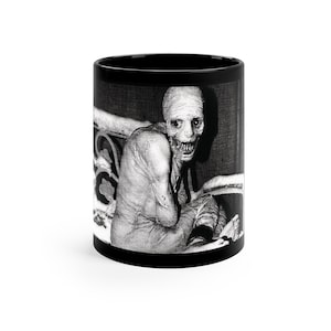 Puede incluir: Taza en blanco y negro con una imagen espeluznante de una criatura con la cara pálida y una amplia sonrisa. La criatura está sentada en una cama.