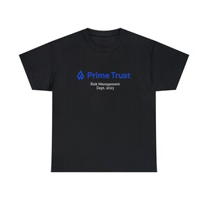 May include: Black t-shirt with the blue text "Prime Trust" and the words "Risk Management Dept. 2023". The shirt has a crew neck and short sleeves. The logo is a blue diamond shape with a flame.