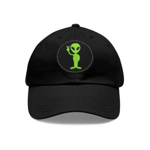 Könnte beinhalten: Schwarze Baseballkappe mit einem grünen Alien-Aufnäher. Der Aufnäher zeigt eine grüne Alien-Figur, die ein Friedenszeichen gibt. Die Kappe besteht aus einem strapazierfähigen Stoff und hat einen gebogenen Schirm. Der Alien-Aufnäher ist kreisförmig mit weißem Rand.