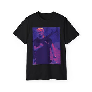 May include: A black t-shirt with a graphic print of a man holding a rifle. The man is wearing glasses and has a beard. The graphic is in shades of purple and pink.