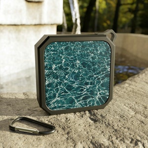 May include: A black portable Bluetooth speaker with a blue and white water-like pattern on the front. The speaker has a built-in carabiner clip.