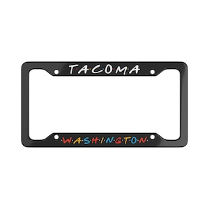 Peut inclure: Cadre de plaque d'immatriculation noir avec le mot "TACOMA" en lettres blanches en haut et "WASHINGTON" en lettres colorées en bas. Le cadre est rectangulaire avec des coins arrondis.