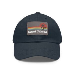 Puede incluir: Una gorra de béisbol azul marino con un parche que presenta una puesta de sol retro, palmeras y el texto "Good Times".