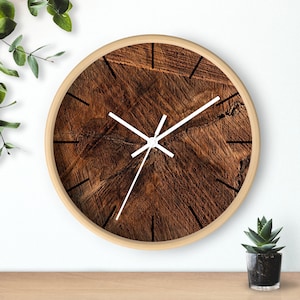 May include: A round wall clock with a light wood frame. The clock face features a detailed wood grain pattern in shades of brown. White hands and black markers indicate the time. The clock is displayed on a wooden surface with greenery in the background.