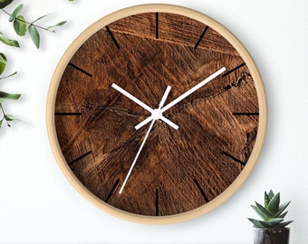 May include: A round wall clock with a light wood frame. The clock face features a detailed wood grain pattern in shades of brown. White hands and black markers indicate the time. The clock is displayed on a wooden surface with greenery in the background.