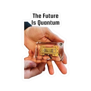 Pode incluir: Um autocolante com o texto "The Future Is Quantum" acima de um bloco retangular transparente contendo uma placa de circuito dourada. O bloco é segurado em mãos abertas.