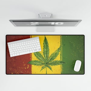Puede incluir: Alfombrilla de escritorio rectangular con un diseño de hoja de cannabis. La alfombrilla tiene un aspecto desgastado con rayas verticales rojas, amarillas y verdes. Un teclado y un ratón blancos están sobre la alfombrilla, con un monitor de ordenador encima.