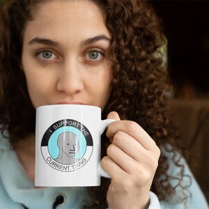 May include: White ceramic mug with a black and white circle design. The circle has a blue background and a gray cartoon face inside. The text "I support the current thing" is written around the circle.