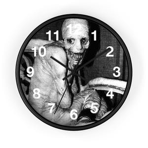 May include: Black and white wall clock with a spooky image of a creature. The clock has a black frame, white numbers, and black hands. The image is a grayscale depiction of a pale, gaunt figure with a menacing expression.