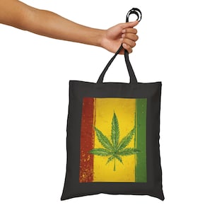 Puede incluir: Bolso tote negro con una hoja de cannabis verde sobre un fondo amarillo con rayas rojas y verdes. El diseño tiene un aspecto vintage y desgastado. El bolso tiene asas negras.