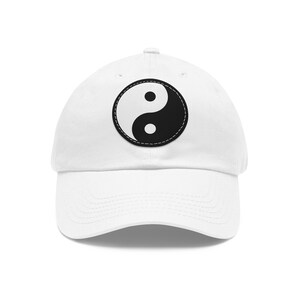 Op de afbeelding: Witte baseballcap met een zwart-wit Yin en Yang symbool. Het symbool is een cirkelvormig ontwerp met een zwart-witte gebogen vorm, gestikt op de voorkant van de cap. De cap heeft een gebogen klep en een stoffen bandsluiting.