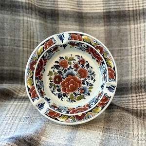 May include: A round, white ceramic ashtray with a floral design. The center features a bouquet of red, blue, and green flowers. The rim is decorated with a repeating pattern of flowers and leaves in red, yellow, and blue.
