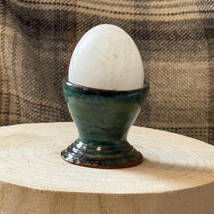 May include: A white, speckled egg sits in a handcrafted, dark green ceramic egg cup. The egg cup has a flared shape with a dark rim and a decorative, ridged base. The cup is placed on a light-colored, wooden surface.