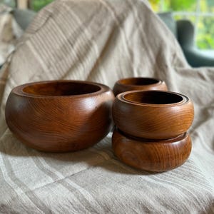 May include: A collection of wooden bowls in varying sizes. The bowls are made of a warm-toned wood, showcasing the natural grain. The largest bowl sits in the foreground, with two smaller bowls stacked behind it, and another bowl to the right.
