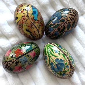 May include: Four decorative, egg-shaped boxes with intricate painted designs. One features yellow birds on a gold background, another has blue flowers on green, and the others have floral patterns in various colors. The boxes are on a white surface.