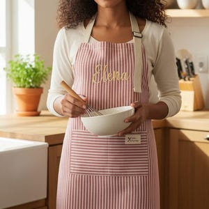 May include: A red and white striped apron with the name "Elena" embroidered in gold script. The apron has a front pocket and is being worn while holding a bowl and whisk. The apron has a small tag that says "X Kitchen Experience".