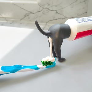 May include: A gray dog-shaped toothpaste tube topper attached to a white and red toothpaste tube, dispensing white toothpaste onto a blue and white toothbrush with green bristles. The topper features a raised tail and four legs.