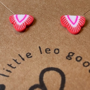 Puede incluir: Un par de pendientes pequeños, rojos y blancos con forma de fresa. Cada pendiente tiene un centro blanco con un contorno rosa y bordes exteriores rojos con líneas blancas. Sobre una superficie marrón, texto "little leo goods" y símbolo del signo zodiacal Leo.