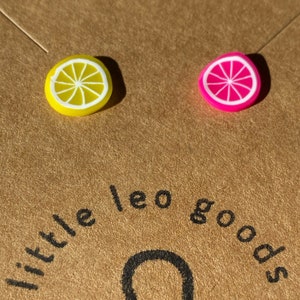 Könnte beinhalten: Ein Paar Ohrringe mit einer gelben Zitronenscheibe und einer rosafarbenen Grapefruitscheibe. Die Ohrringe befinden sich auf einer braunen Oberfläche mit dem Text "little leo goods" und dem astrologischen Symbol für Löwe.