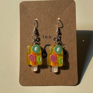 Pode incluir: Um par de brincos de picolé amarelo com um design de kiwi, morango e melancia. Os brincos estão em um cartão marrom com o texto "leo"
