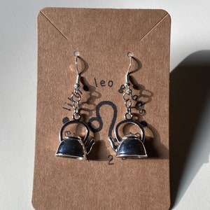 May include: Silver teapot dangle earrings on a brown card with the text "Leo Goods" and a Leo zodiac symbol.