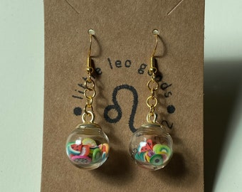 May include: Gold dangle earrings with a clear glass orb filled with colorful fruit slices. The earrings are on a brown card with the text "little leo goods" and a zodiac symbol.