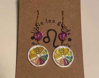 May include: A pair of gold earrings with a pink bead and a colorful lemon slice charm. The earrings are labeled "Little Leo Goods" with the astrological symbol for Leo.