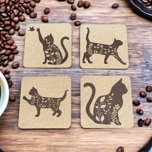 May include: Set of four brown cork coasters with black silhouette images of cats with floral designs. The cats are in different poses, including sitting, standing, and lying down.