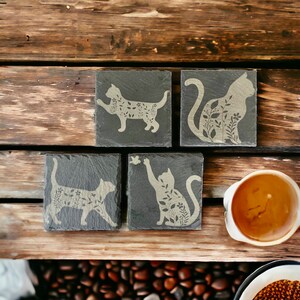 May include: Four black slate coasters with a white silhouette of a cat with floral details on each coaster.