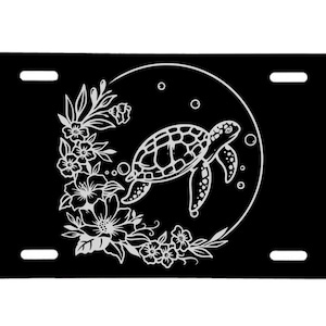 May include: Black license plate featuring a white sea turtle design within a circle, surrounded by floral and botanical elements. The design includes a detailed turtle illustration and decorative flowers.