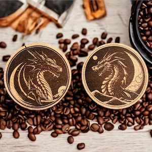 May include: Two round wooden coasters with a dark brown background and a light brown design of a dragon with a crescent moon. The coasters are surrounded by coffee beans.